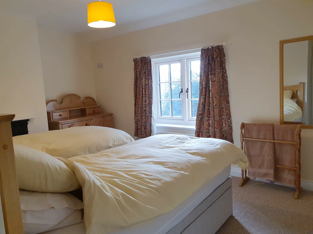 Two twin beds with beige bedding, window with patterned curtains, wooden dresser, mirror, and yellow pendant light