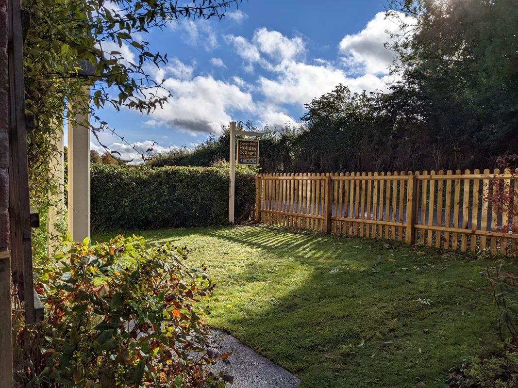 Sunny garden with wooden fence, gravel path, and 'Holiday Cottages' sign. Lush greenery and clear sky.