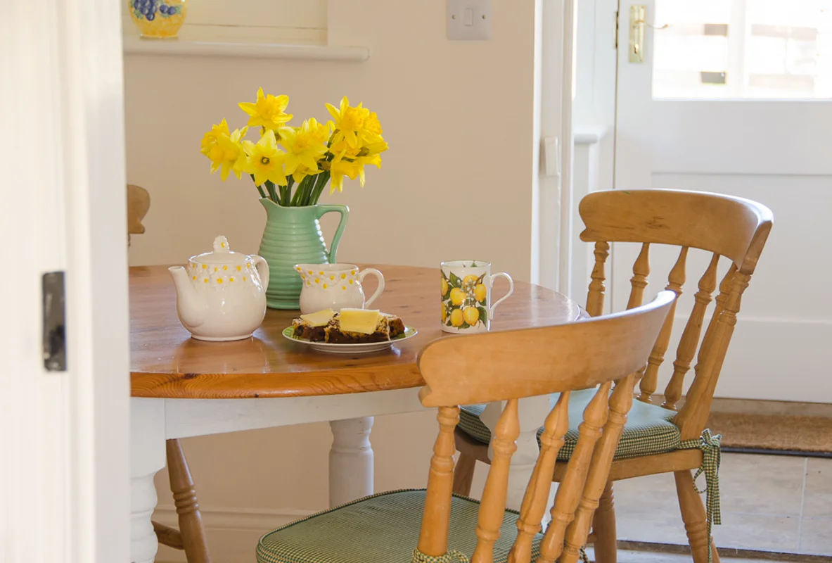 Wooden table with yellow daffodils, teapot, lemon mug, and cake in sunny cottage setting.
