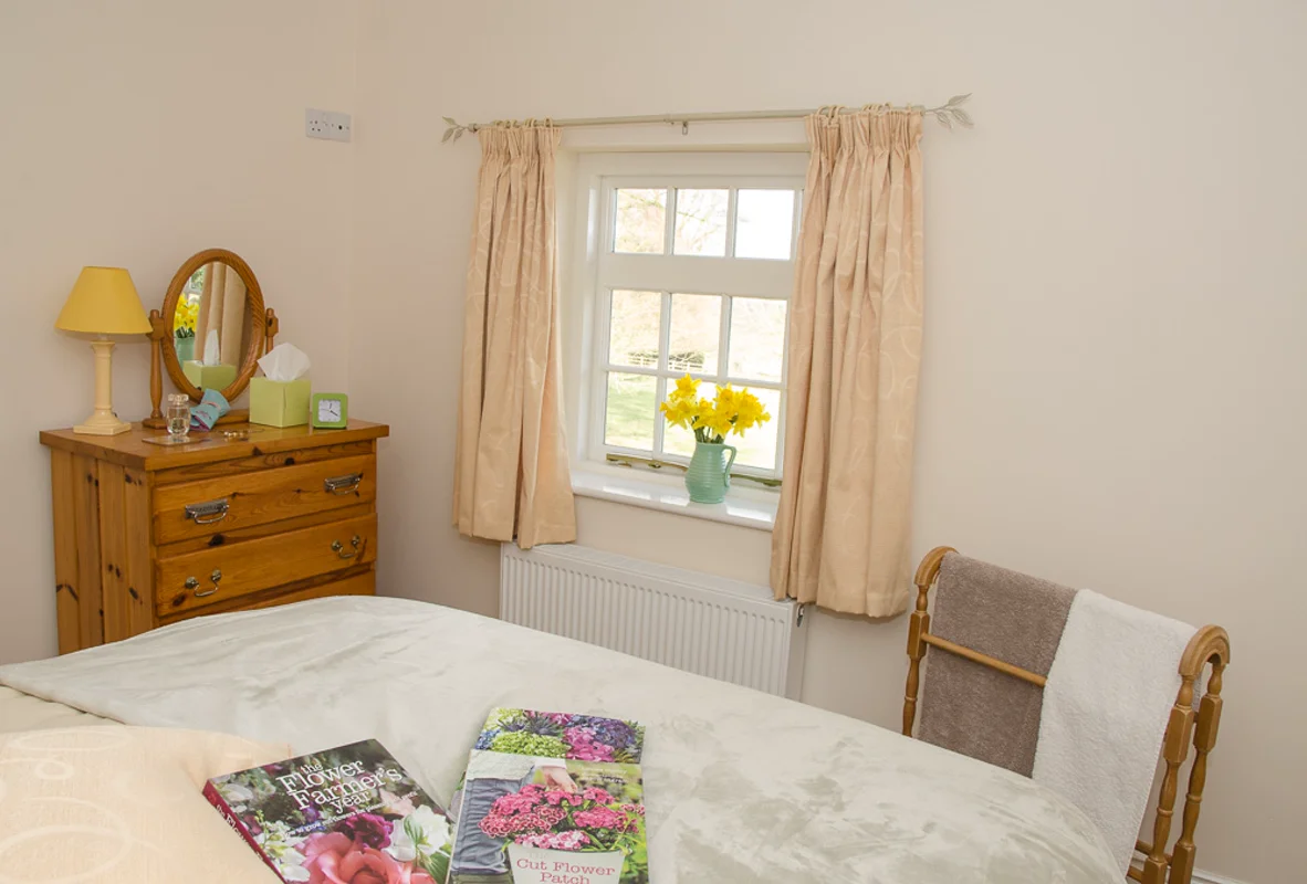 Cottage bedroom with beige curtains, yellow flowers on windowsill, wooden dresser with lamp and mirror, bed with books, chair with towels.