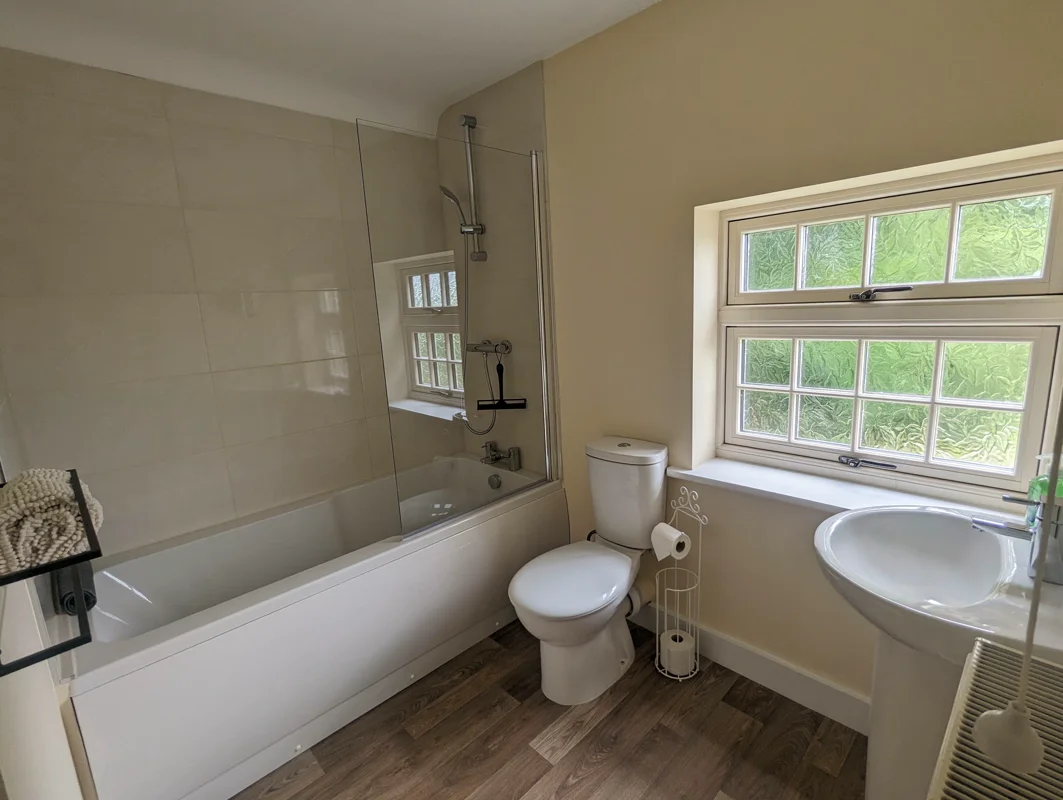 Bright bathroom with white tub, shower, toilet, sink, and large window overlooking greenery