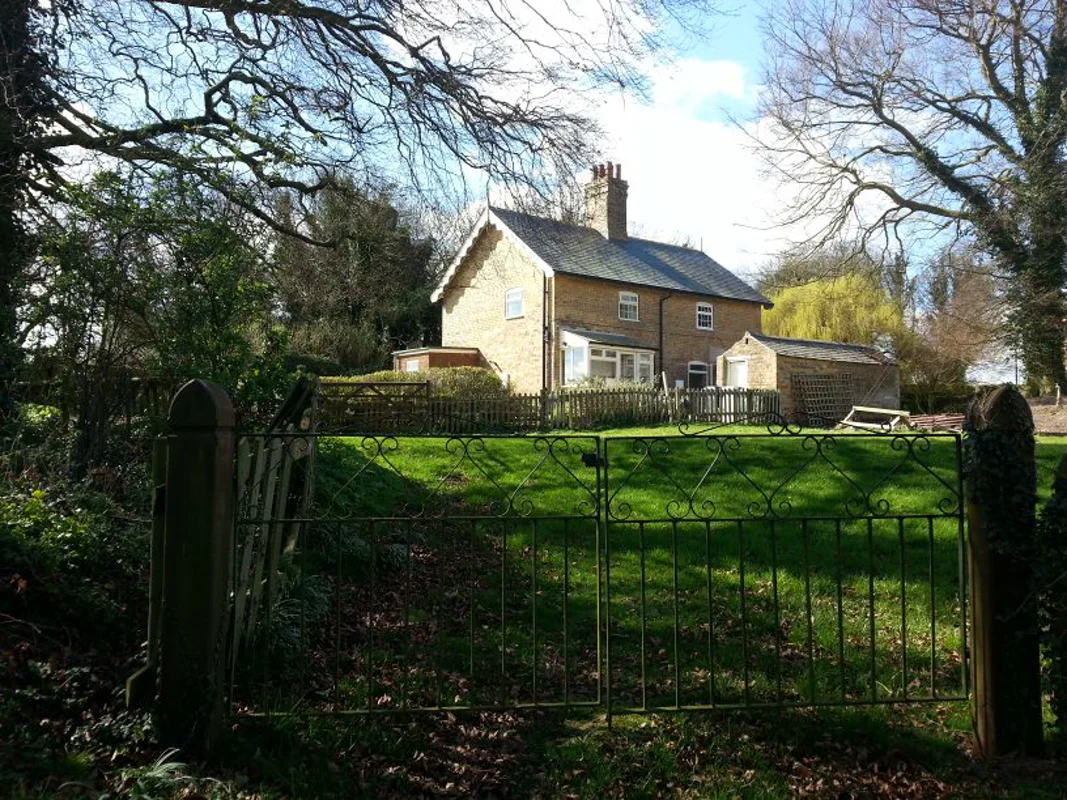 Stone cottage with garden, iron gate, and trees in sunny setting.