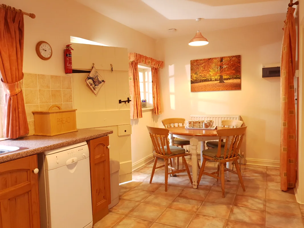 Warm-toned cottage kitchen with wooden dining table, tiled floor, autumn artwork, soft orange curtains, and kitchen appliances.