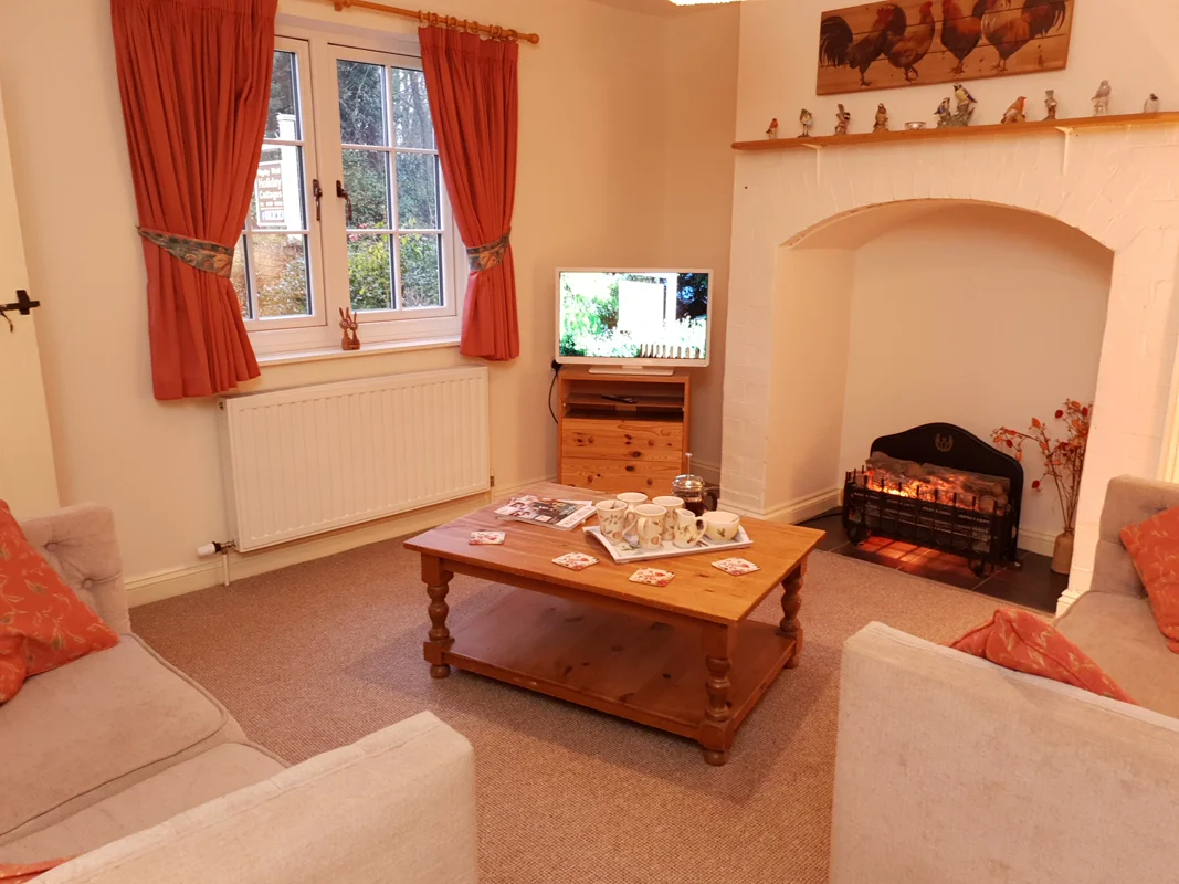 Cottage living room with cream sofas, red curtains, wooden coffee table, fireplace, TV, and garden view.