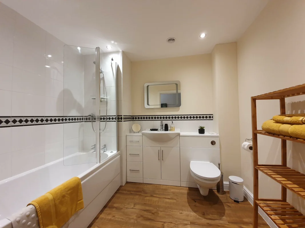 Clean bathroom: white tiles, bathtub, glass shower, yellow towels, wooden shelves, sink, toilet. Warm lighting.