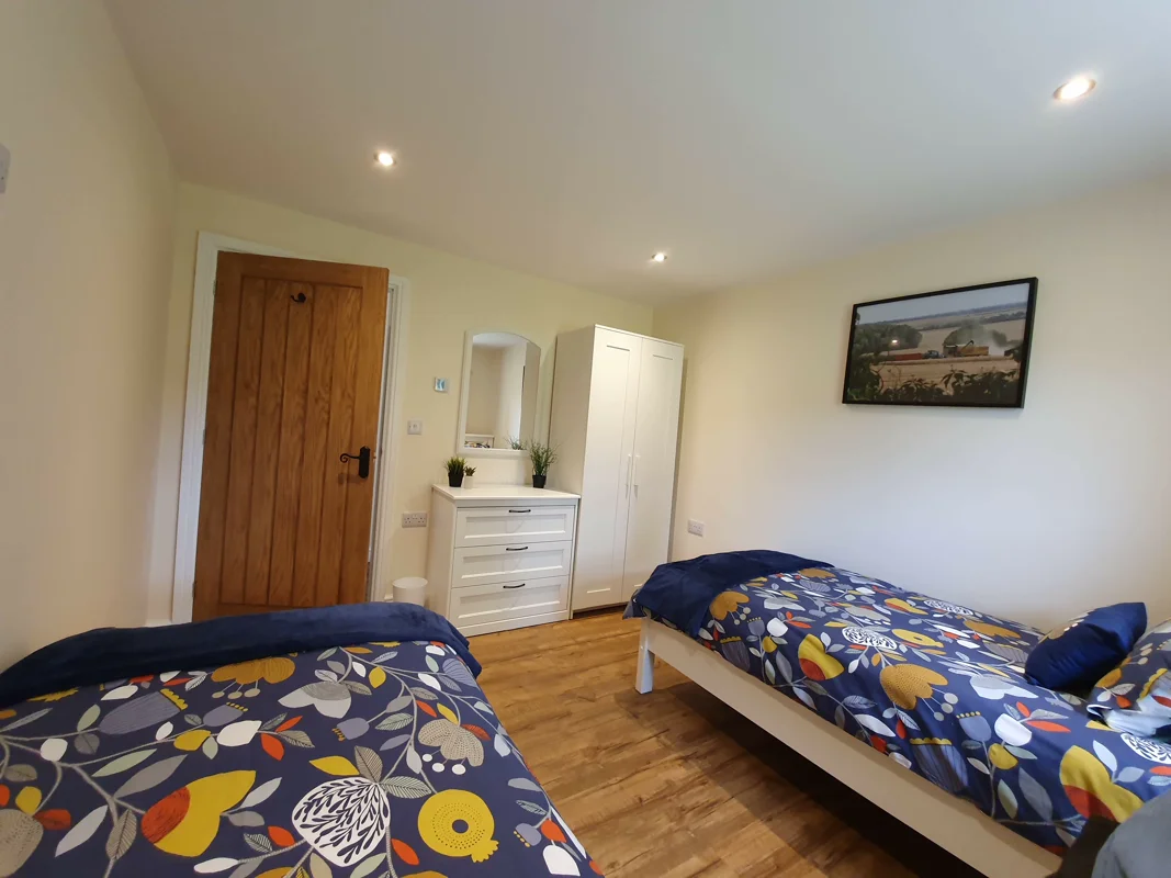 Two single beds with colorful floral duvets, wooden door, white dresser, and wardrobe.