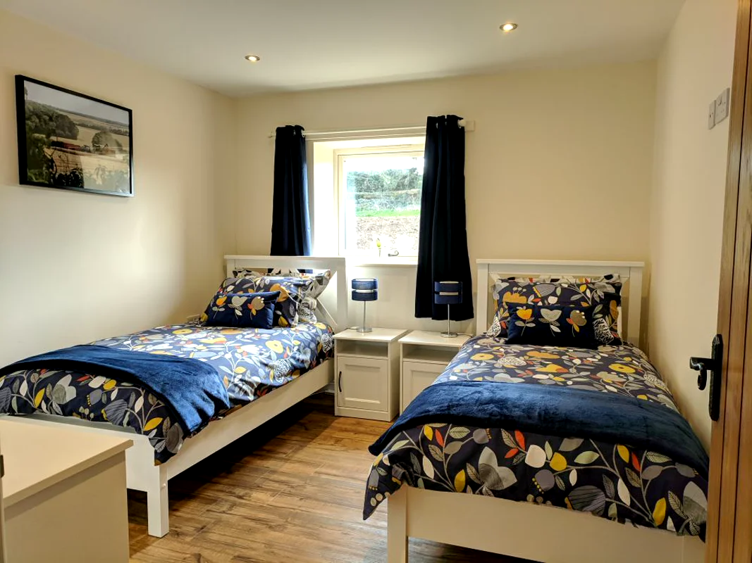 Two single beds with floral blue bedding, white nightstands, dark curtains. Sunlit holiday cottage bedroom.