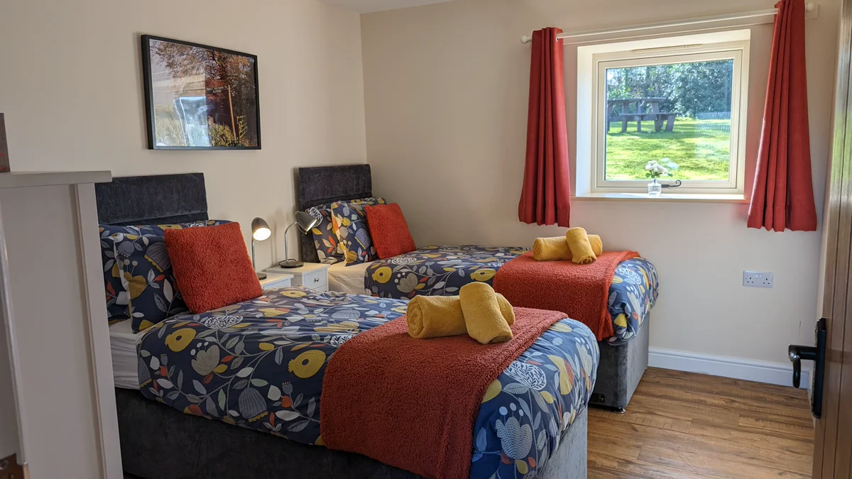 Brightly decorated twin bedroom with colorful bedding, red curtains, and garden view through window.