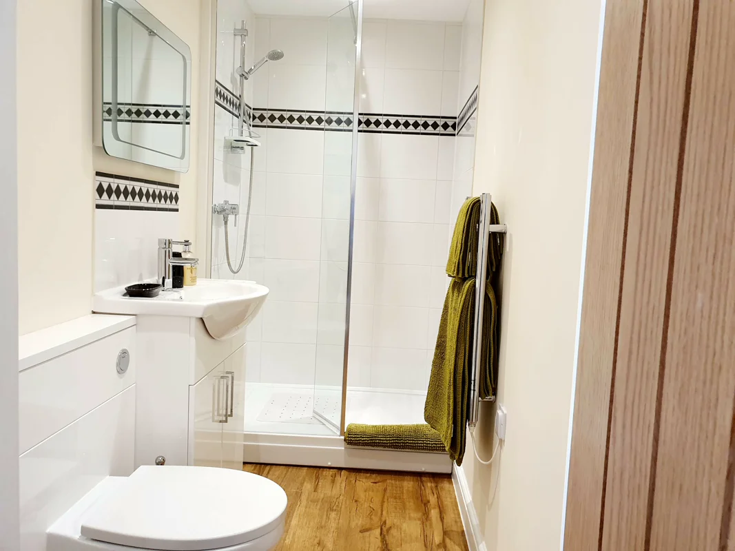 Clean bathroom with shower, sink, toilet, green towels, and wooden flooring