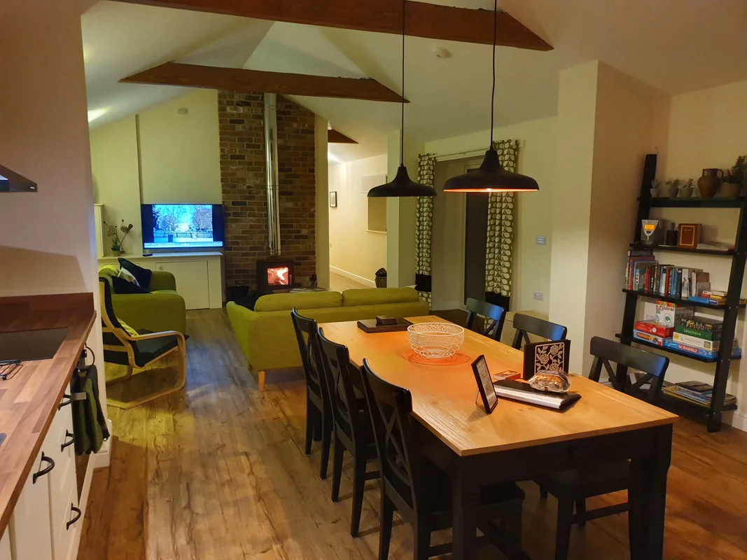 Warmly lit cottage interior with wooden dining table, green sofas, brick fireplace, TV. Modern kitchen visible.