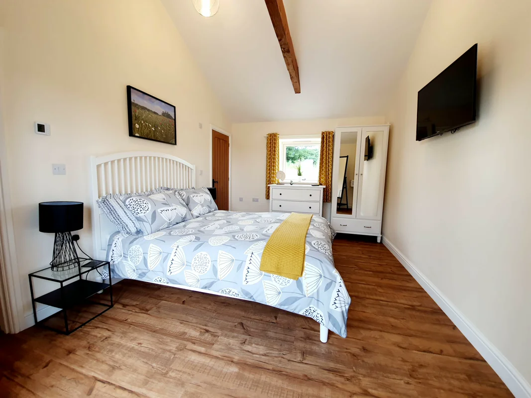 Light-filled cottage bedroom with white bed, patterned bedding, yellow throw, wooden flooring, and modern furniture.